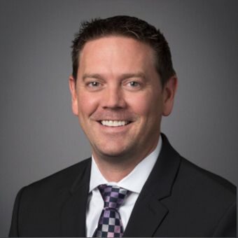 A man in a black suit and patterned tie smiles against a gray background, exuding confidence like a seasoned land surveyor ready to tackle any terrain.